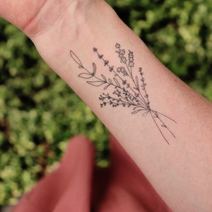 May include: A black line art tattoo of a floral bouquet on a person's forearm. The bouquet includes sprigs of lavender and other small flowers, tied together with a simple line. The background is blurred green foliage and a red-orange fabric.
