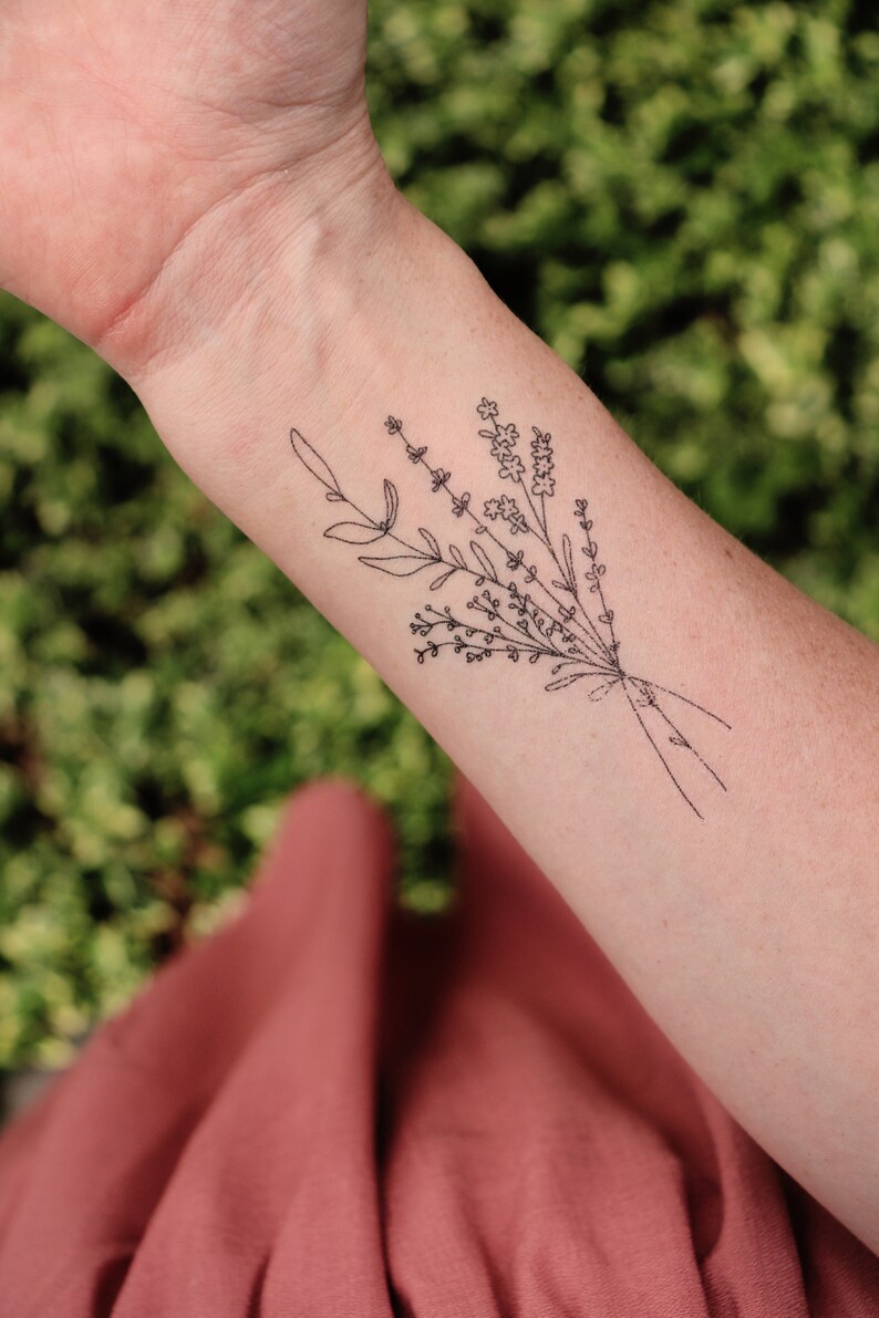May include: A black line art tattoo of a floral bouquet on a person's forearm. The bouquet includes sprigs of lavender and other small flowers, tied together with a simple line. The background is blurred green foliage and a red-orange fabric.