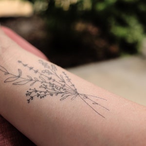 May include: A black and white floral tattoo design on the forearm. The tattoo features a delicate bouquet of leaves and small flowers, tied together with thin lines. The skin tone is light.
