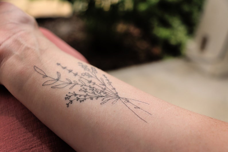 May include: A black and white floral tattoo design on the forearm. The tattoo features a delicate bouquet of leaves and small flowers, tied together with thin lines. The skin tone is light.