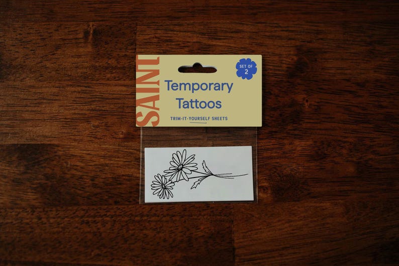 Pu&ograve; includere: Un pacchetto di tatuaggi temporanei con un disegno a margherita. La confezione recita "SAINT Temporary Tattoos" e "TRIM-IT-YOURSELF SHEETS". Il set include due fogli di tatuaggi.