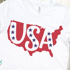 May include: White t-shirt with a red outline of the United States with the word "USA" in white letters and blue stars inside the letters and outline.