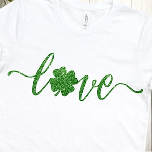 Puede incluir: Camiseta blanca con la palabra "love" escrita en letras de purpurina verde con un tr&eacute;bol de purpurina verde en el medio de la palabra.