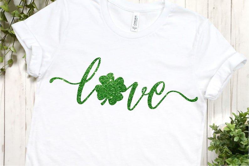 Puede incluir: Camiseta blanca con la palabra "love" escrita en letras de purpurina verde con un tr&eacute;bol de purpurina verde en el medio de la palabra.