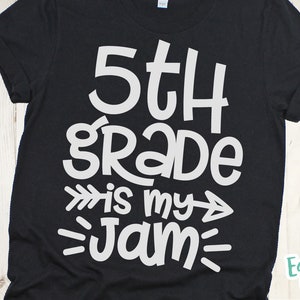 Op de afbeelding: Zwart T-shirt met witte tekst die "5th grade is my jam" luidt met pijlen en lijnen aan beide kanten van de tekst.