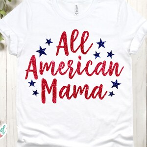 Puede incluir: Camiseta blanca con la frase "All American Mama" en rojo brillante, rodeada de estrellas azules. La camiseta se muestra sobre un fondo de madera clara con follaje verde.