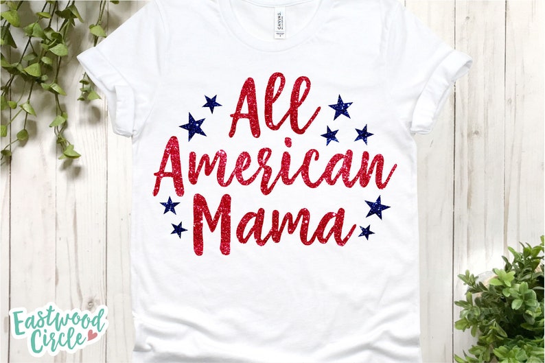 Puede incluir: Camiseta blanca con la frase "All American Mama" en rojo brillante, rodeada de estrellas azules. La camiseta se muestra sobre un fondo de madera clara con follaje verde.