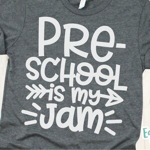 Op de afbeelding: Een donkergrijs T-shirt met witte tekst die "Pre-school is my jam" luidt, met een witte pijl die naar rechts en links van de tekst wijst.