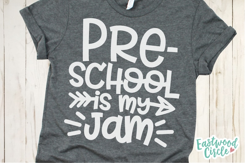 Op de afbeelding: Een donkergrijs T-shirt met witte tekst die "Pre-school is my jam" luidt, met een witte pijl die naar rechts en links van de tekst wijst.