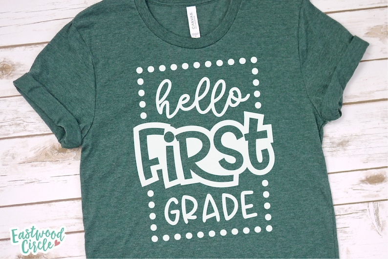 May include: A green t-shirt with the words "hello FIRST GRADE" in white, framed by a dotted border. The shirt has rolled-up sleeves and a crew neck. The brand "Eastwood Circle" is printed on the bottom left.