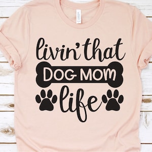 May include: A pink t-shirt with the text "livin' that DOG MOM life" in black lettering. The text is surrounded by a black bone and two black paw prints.