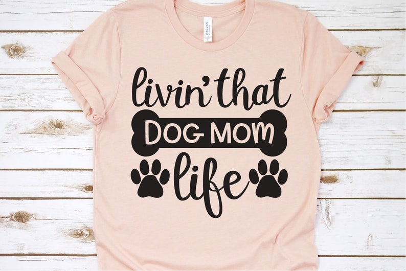 May include: A pink t-shirt with the text "livin' that DOG MOM life" in black lettering. The text is surrounded by a black bone and two black paw prints.