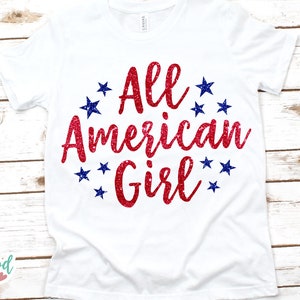Puede incluir: Camiseta blanca con el texto "All American Girl" en letras rojas brillantes, rodeada de estrellas azules. La camiseta se muestra sobre una superficie de madera clara.