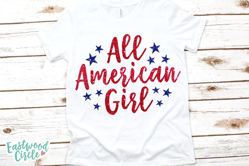 Puede incluir: Camiseta blanca con el texto "All American Girl" en letras rojas brillantes, rodeada de estrellas azules. La camiseta se muestra sobre una superficie de madera clara.
