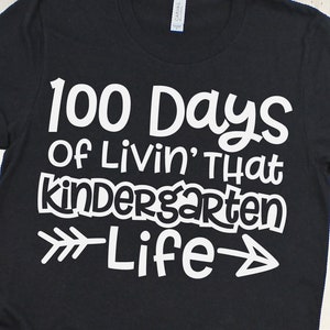 Puede incluir: Camiseta negra con texto blanco que dice "100 Days of Livin' That Kindergarten Life" con una flecha blanca apuntando hacia la derecha y la izquierda.
