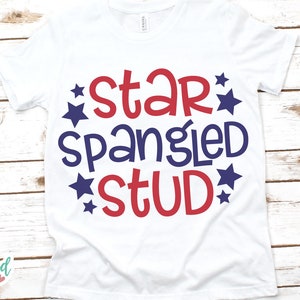 Puede incluir: Camiseta blanca con la frase "Star Spangled Stud" en rojo y azul, rodeada de estrellas azules. La camiseta est&aacute; sobre una superficie de madera blanca. El texto est&aacute; en una fuente audaz.