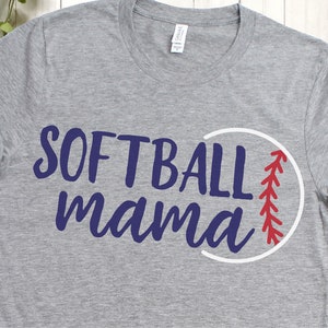May include: A gray t-shirt with the words "SOFTBALL mama" in navy blue script. A white circle with red stitching, resembling a softball, is next to the text. The shirt has rolled-up sleeves.