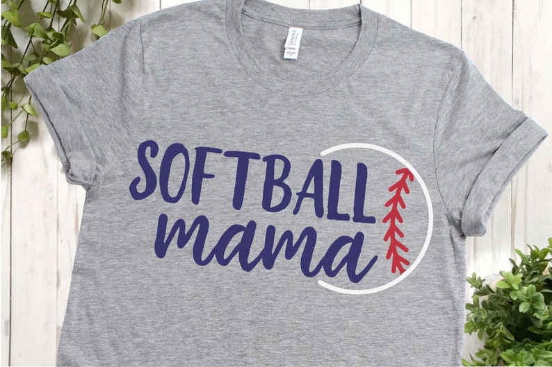May include: A gray t-shirt with the words "SOFTBALL mama" in navy blue script. A white circle with red stitching, resembling a softball, is next to the text. The shirt has rolled-up sleeves.
