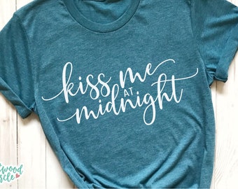 Può includere: T-shirt color verde acqua con la scritta bianca "kiss me at midnight" in corsivo. La maglietta ha le maniche arrotolate e lo scollo tondo. Il logo Eastwood Circle è visibile in basso a sinistra.