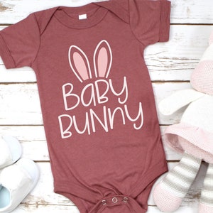 May include: A maroon baby onesie with white text that reads "Baby Bunny" and pink bunny ears. The onesie is lying on a white wooden surface with a white and pink stuffed unicorn toy.