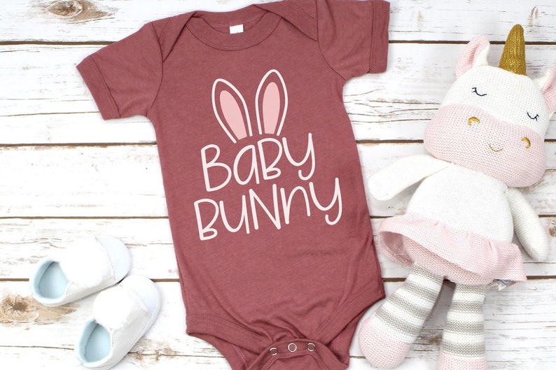 May include: A maroon baby onesie with white text that reads "Baby Bunny" and pink bunny ears. The onesie is lying on a white wooden surface with a white and pink stuffed unicorn toy.