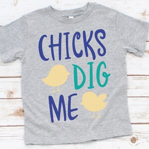 May include: A gray t-shirt with the text "CHICKS DIG ME" in blue and teal lettering. Two yellow chicks are on either side of the text.