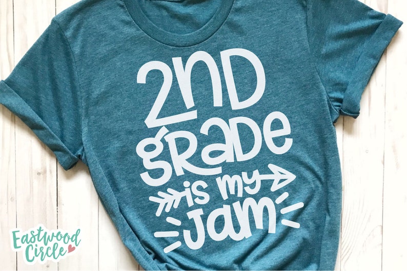 Op de afbeelding: Tealblauw T-shirt met witte tekst die "2nd grade is my jam" zegt met een pijl die naar rechts wijst.