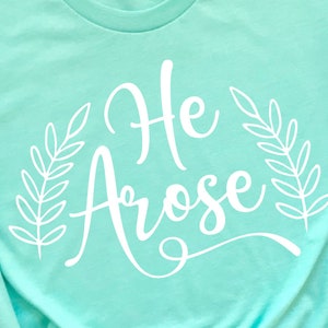 May include: A turquoise t-shirt with the text "He Arose" in white cursive lettering, surrounded by two white leafy branches.