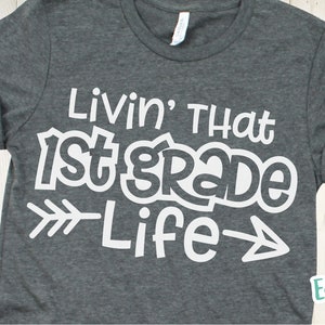Puede incluir: Una camiseta gris oscura con texto blanco que dice "Livin' That 1st Grade Life" con flechas a cada lado de la palabra "Life".