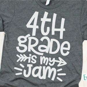 Op de afbeelding: Een grijs T-shirt met witte tekst die "4th grade is my jam" zegt met pijlen aan beide kanten van "is my".