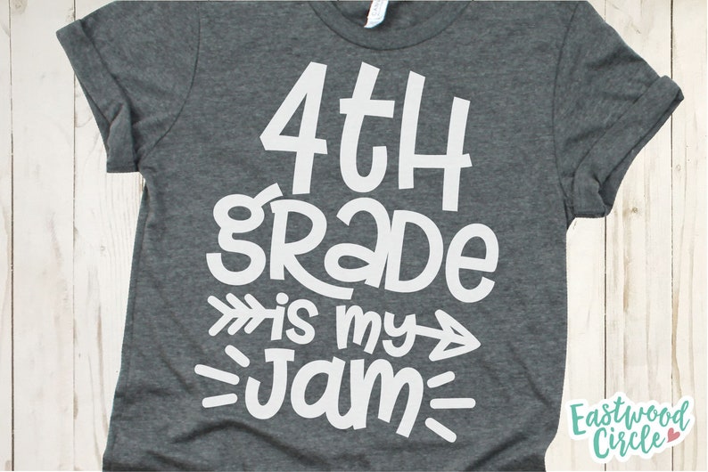 Op de afbeelding: Een grijs T-shirt met witte tekst die "4th grade is my jam" zegt met pijlen aan beide kanten van "is my".