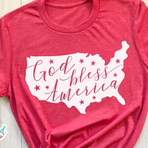 Puede incluir: Camiseta roja con mangas enrolladas. Presenta un contorno blanco de los Estados Unidos con la frase "God bless America" en cursiva y estrellas.