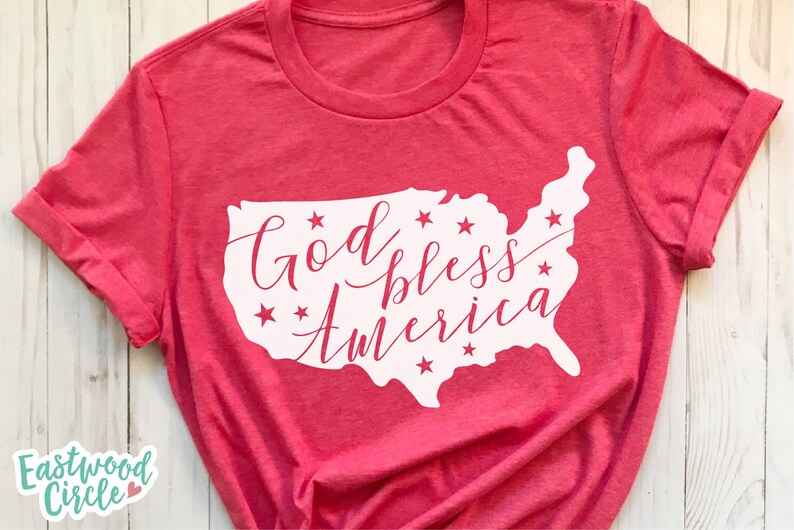 Puede incluir: Camiseta roja con mangas enrolladas. Presenta un contorno blanco de los Estados Unidos con la frase "God bless America" en cursiva y estrellas.