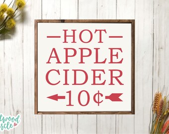 Puede incluir: Un letrero de madera enmarcado con las palabras "HOT APPLE CIDER" en rojo, con una flecha que apunta a la izquierda y el texto "10¢". El letrero está sobre un fondo blanco con un marco de madera.