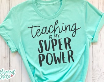 Puede incluir: Una camiseta turquesa con el texto "Teaching is my superpower" en letras negras.