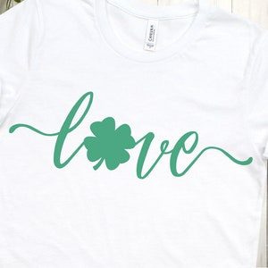 Puede incluir: Camiseta blanca con la palabra "love" escrita en cursiva con un tr&eacute;bol de cuatro hojas verde en el centro de la palabra.