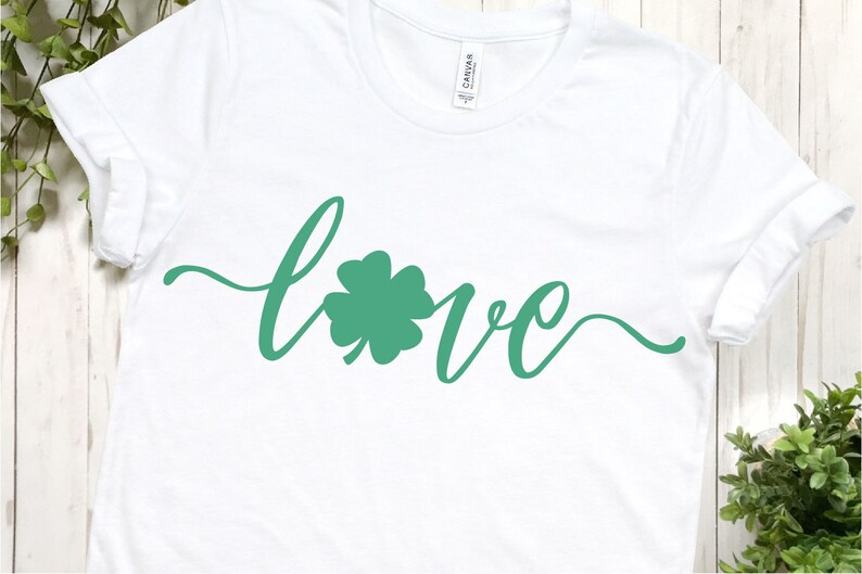 Puede incluir: Camiseta blanca con la palabra "love" escrita en cursiva con un tr&eacute;bol de cuatro hojas verde en el centro de la palabra.