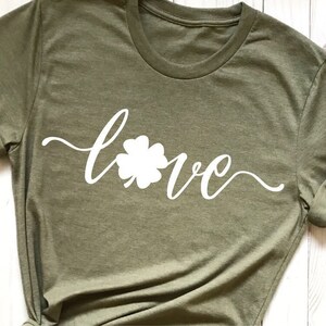 Puede incluir: Una camiseta verde con un tr&eacute;bol de cuatro hojas blanco y la palabra "love" escrita en cursiva.