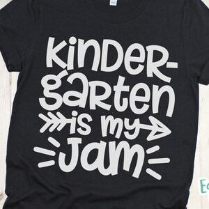 Op de afbeelding: Zwart T-shirt met witte tekst die "Kindergarten is my jam" zegt met pijlen aan beide kanten van het woord "my".