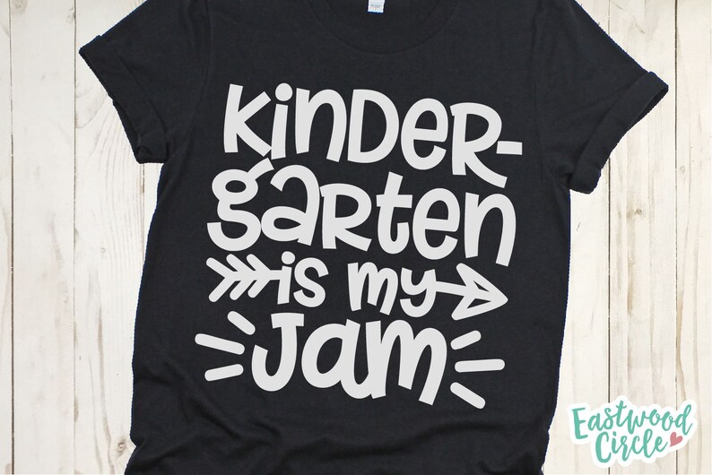 Op de afbeelding: Zwart T-shirt met witte tekst die "Kindergarten is my jam" zegt met pijlen aan beide kanten van het woord "my".