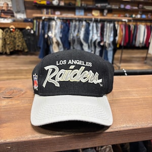 Puede incluir: Gorra de béisbol negra y gris con el texto "LOS ANGELES Raiders" en letras blancas y doradas. La gorra tiene una visera gris y un logotipo del equipo. El sombrero está hecho de un material similar a la lana.