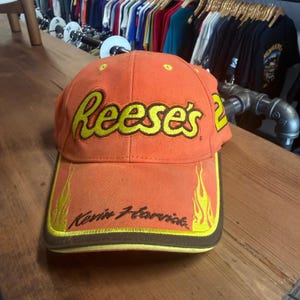 May include: Orange baseball cap with the Reese's logo in yellow. The brim has yellow flames and the name "Kevin Harvick" in black script. The cap has a brown trim and the number 2 on the side.