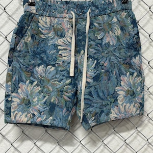 May include: Shorts featuring a woven tapestry fabric with a dense floral pattern in shades of blue, teal, and soft pink. The garment includes an elasticized waistband with a long, flat, off-white drawstring cord and two side pockets.