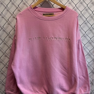Peut inclure: Un sweat-shirt rose à col rond avec le texte "b.u.m. equipment" brodé sur la poitrine. Le sweat-shirt est suspendu à un cintre en bois sur un fond de clôture en treillis métallique.