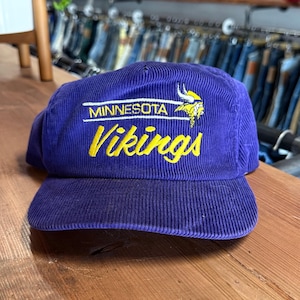 May include: A purple corduroy baseball cap featuring the embroidered text Minnesota Vikings in yellow and white, accompanied by a small yellow and white Viking helmet logo. The cap has a structured crown and a flat brim, showcasing a textured fabric.