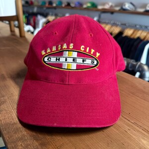 Peut inclure: Casquette de baseball rouge avec l'inscription brodée "KANSAS CITY CHIEFS" en jaune et blanc. La casquette a une visière incurvée et une couronne structurée. Le logo est de forme ovale avec une bande blanche et jaune.
