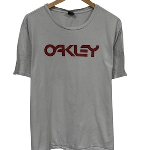 May include: A light gray t-shirt with the brand name "OAKLEY" printed in a dark red color across the chest. The shirt has short sleeves and a crew neck, hanging on a wooden hanger.