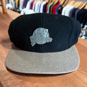 Puede incluir: Una gorra de béisbol Planet Hollywood negra y gris. La gorra tiene una corona negra y una visera gris. Un logotipo plateado de Planet Hollywood está centrado en la parte delantera.