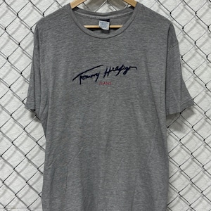 May include: A gray Tommy Hilfiger Jeans t-shirt hanging on a wooden hanger. The shirt features the Tommy Hilfiger logo in navy blue script across the chest. The word "JEANS" is printed below the logo in navy blue.