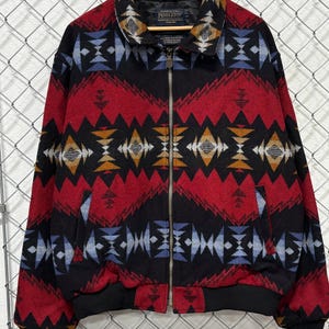 May include: A Pendleton zip-up jacket featuring a bold geometric pattern in red, black, blue, and gold. The jacket has a fold-down collar, a full-length metal zipper, two side pockets, and black ribbed cuffs and hem. The Pendleton brand label is visible.